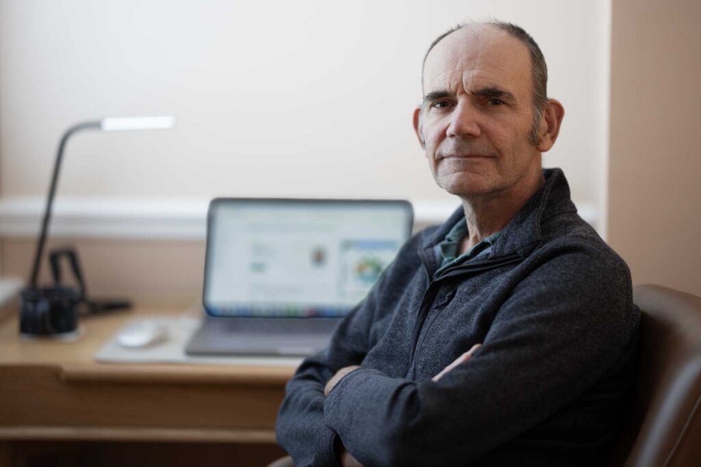 Man sitting indoors with folded arms beside a desk, with a laptop and lamp blurred in the background.