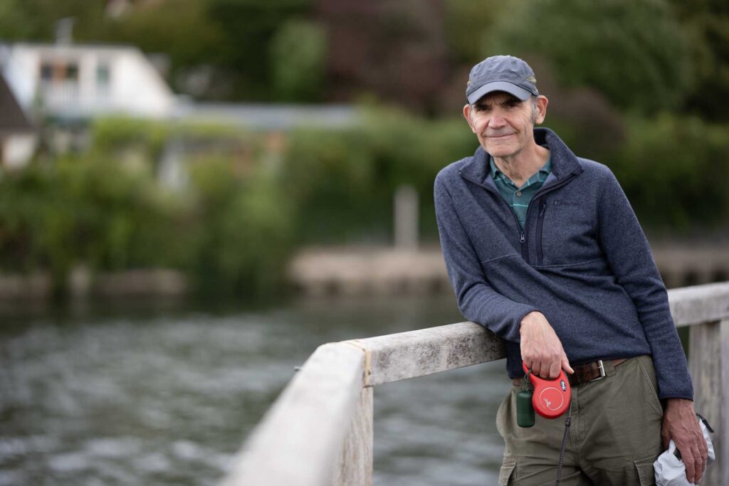 Man wearing a cap leaning against a wooden railing beside water, with trees and buildings softly blurred behind him.