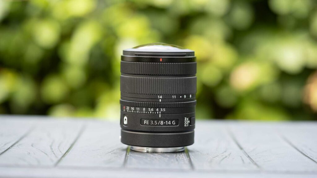 Sony FE 8-14mm F3.5 Fisheye G set to 14mm, standing on a grey table against blurred green foliage.