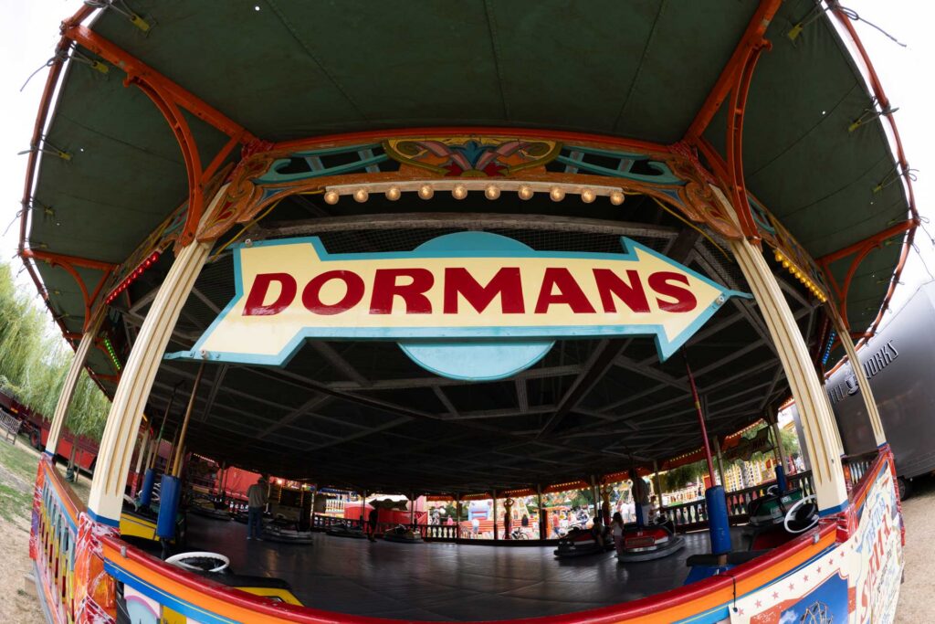 Fisheye view of a brightly painted dodgems pavilion with a large red “DORMANS” sign above the bumper-car floor.