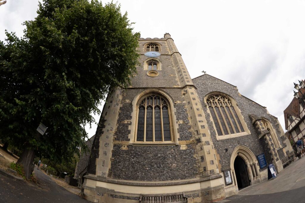 Low-angle fisheye view of a flint church with tall arched windows and tower, framed by a large leafy tree and cloudy sky.