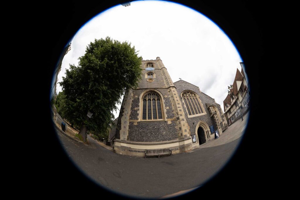 ircular fisheye view of a flint church, tree and neighbouring buildings contained within a round image surrounded by black.