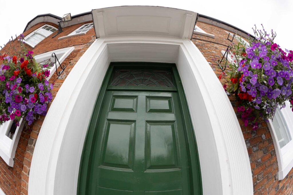 Low-angle fisheye view of a dark green front door in a red-brick building, flanked by hanging baskets of purple, pink and red flowers.
