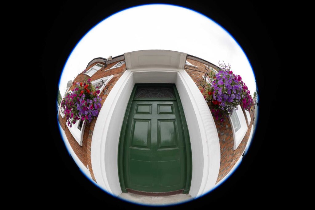 Circular fisheye view of a green front door and flower-filled hanging baskets, surrounded by a broad black border.