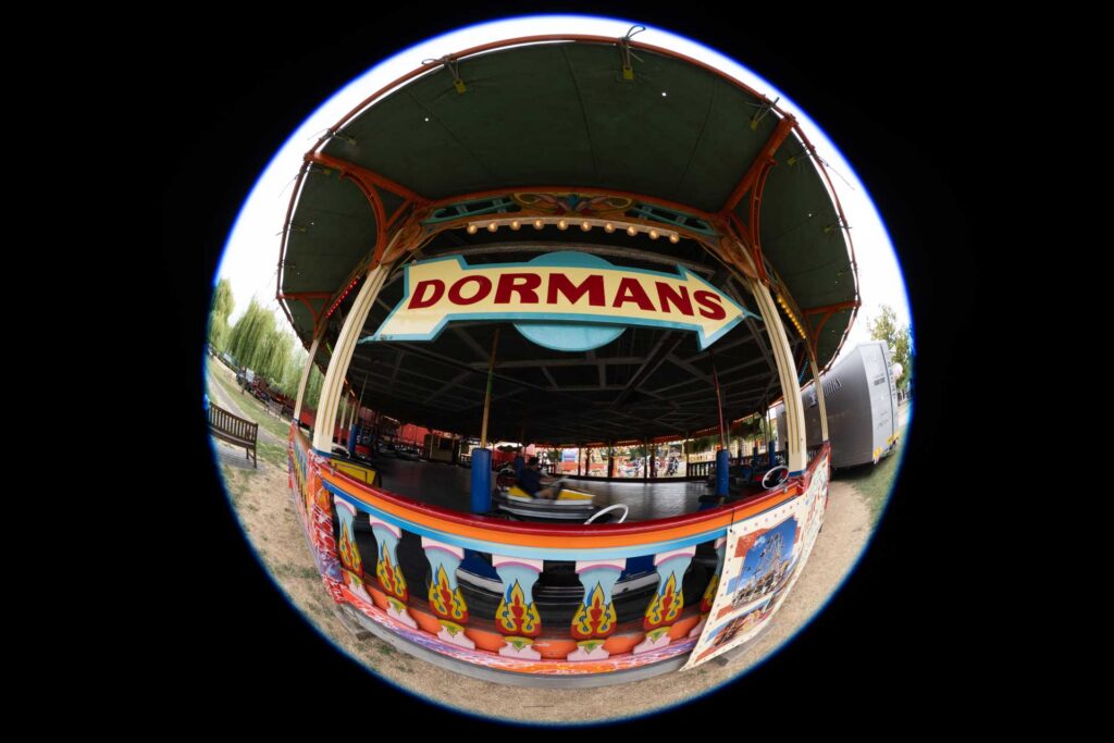 Circular fisheye view of the DORMANS dodgems pavilion with bumper cars moving inside, surrounded by a black frame.