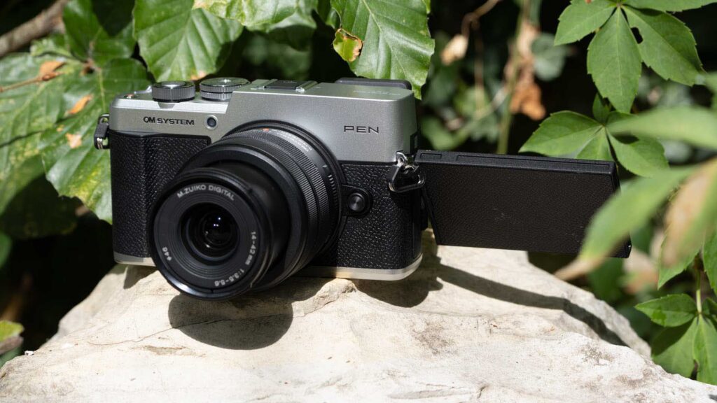 Front three-quarter view of the OM System PEN and 14–42mm lens on a rock, with its vari-angle touchscreen opened to the right.