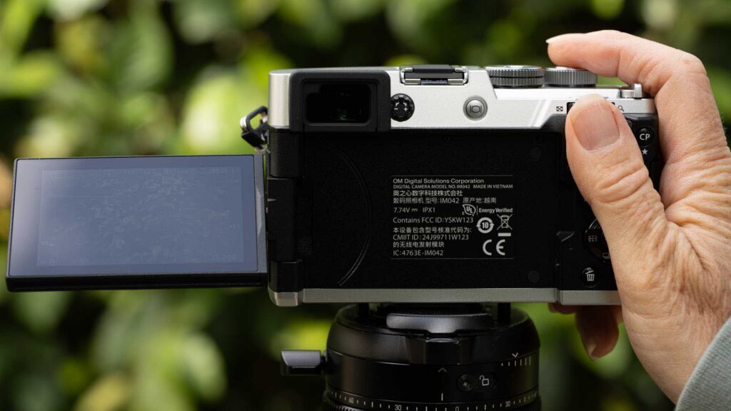 Rear view of the OM System PEN on a tripod, with its touchscreen swung out to the left and a hand resting on the top controls.