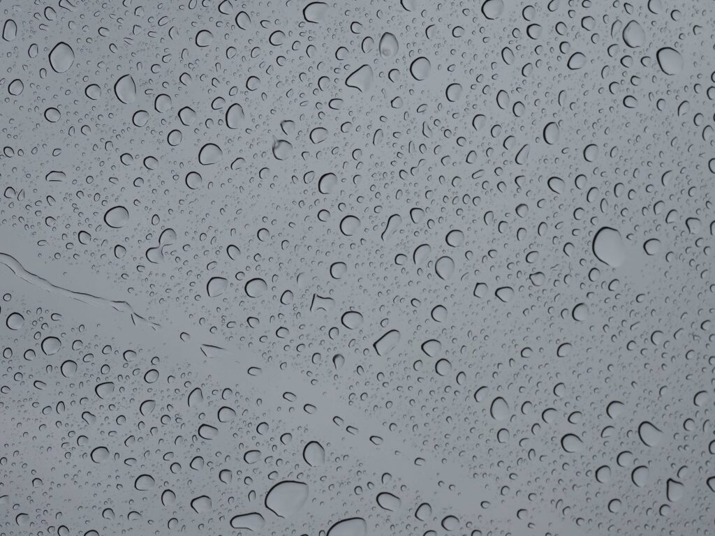 Raindrops of varying sizes covering a glass surface against a pale grey sky.