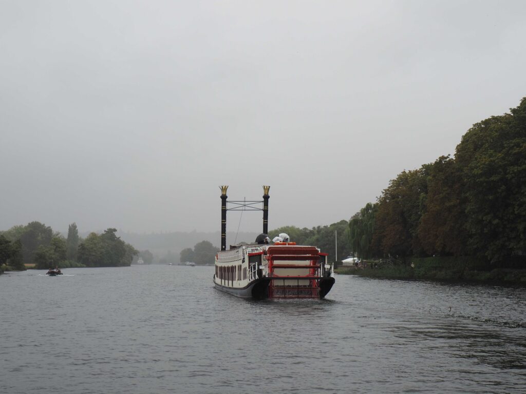 Paddle steamer travelling along a broad river in rainy, misty weather, with tree-lined banks fading into the distance.