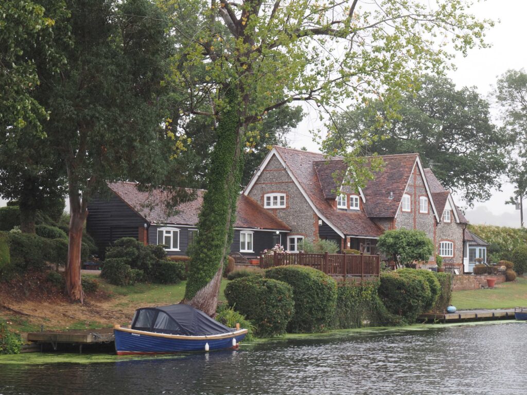 Riverside house with brick, flint and black timber walls, surrounded by mature trees and shrubs, with a small blue boat moored in front.