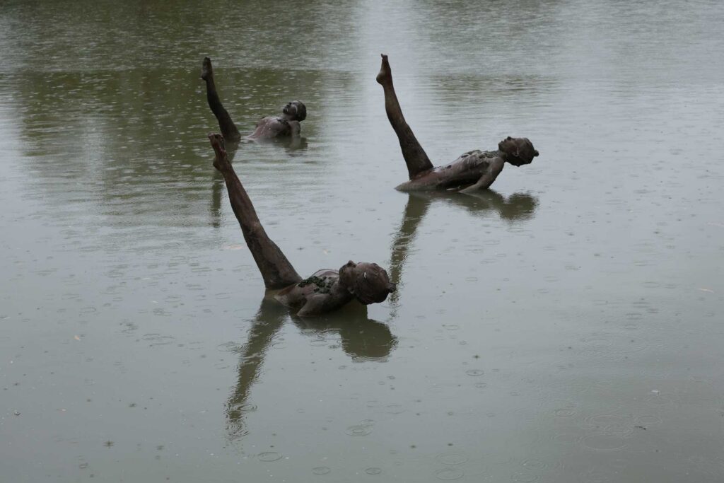 Three bronze human figures partially submerged in a rain-dappled pond, each posed with one leg raised vertically.
