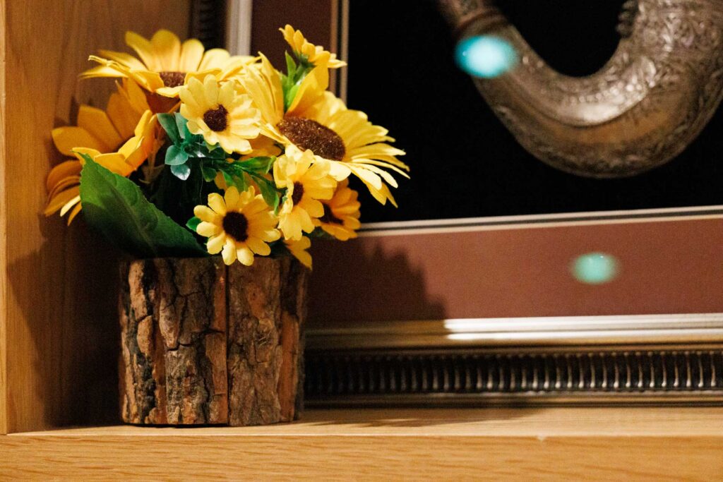 Close view of yellow artificial flowers in a bark-covered pot, lit warmly against framed wall decoration.