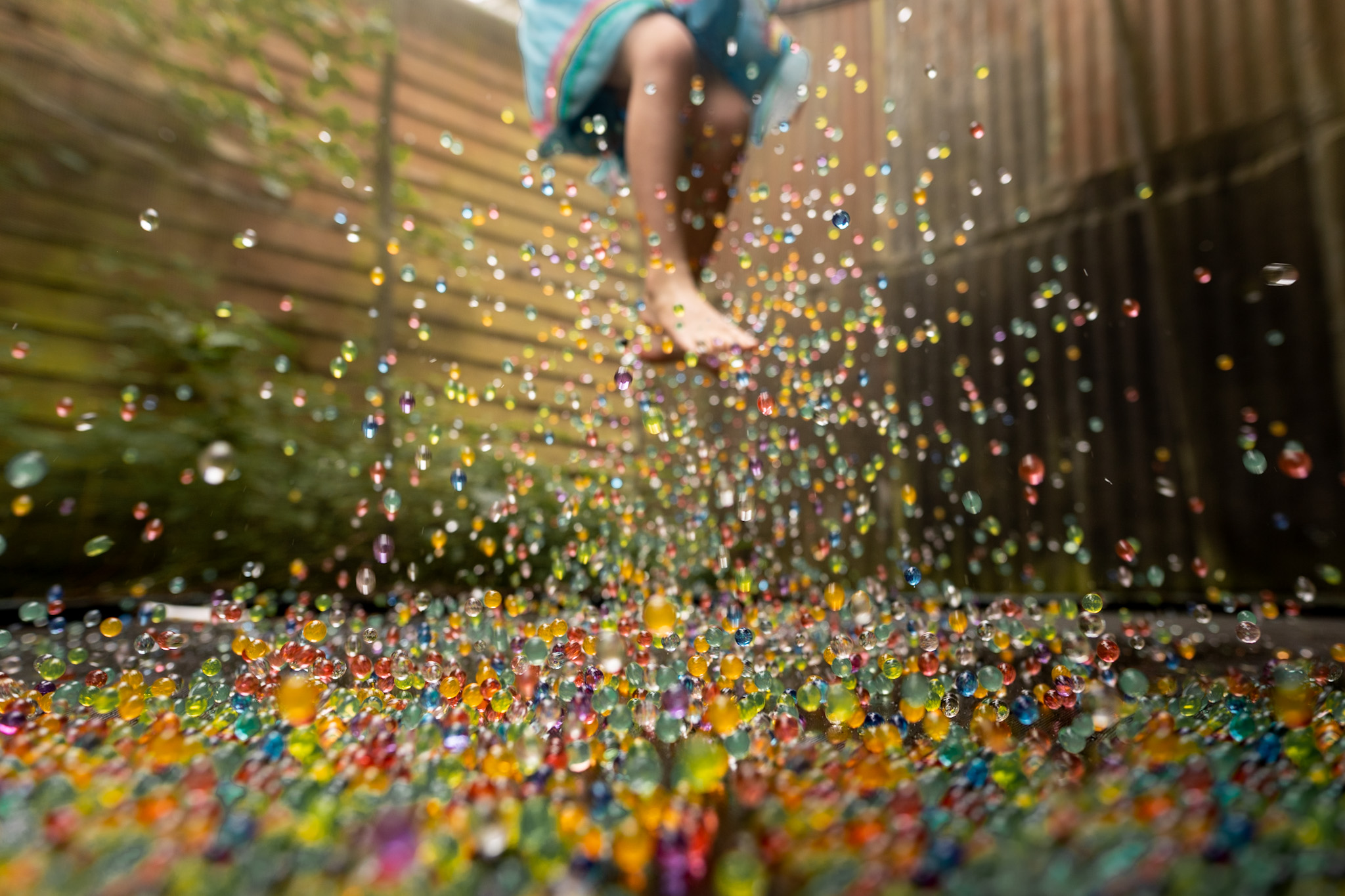 Hundreds of colourful translucent water beads bounce through the air beneath a child's bare feet in a garden.