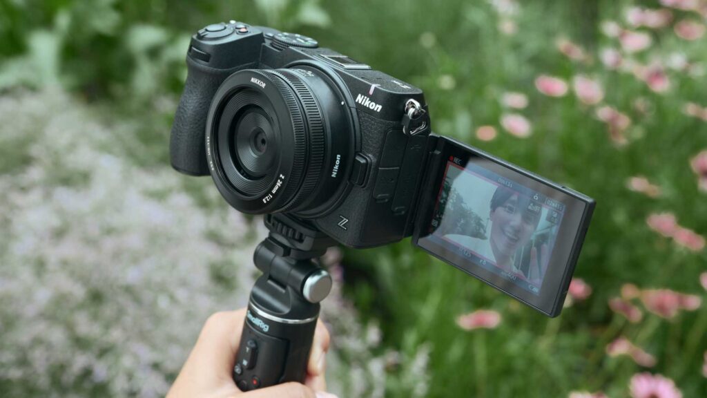 A black Nikon Z5IIC mounted on a handheld grip outdoors, with its screen swung out to the side.