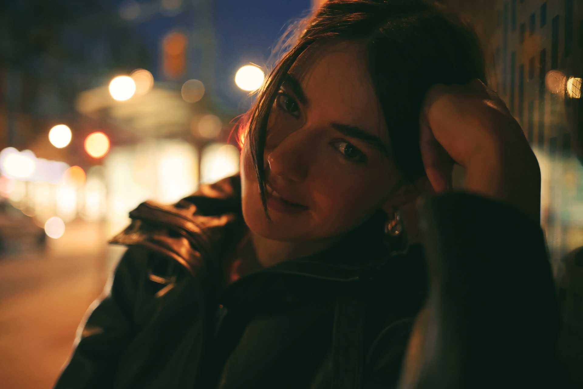 A woman in a dark jacket rests her head on her hand on a city street at night, with blurred lights behind her.