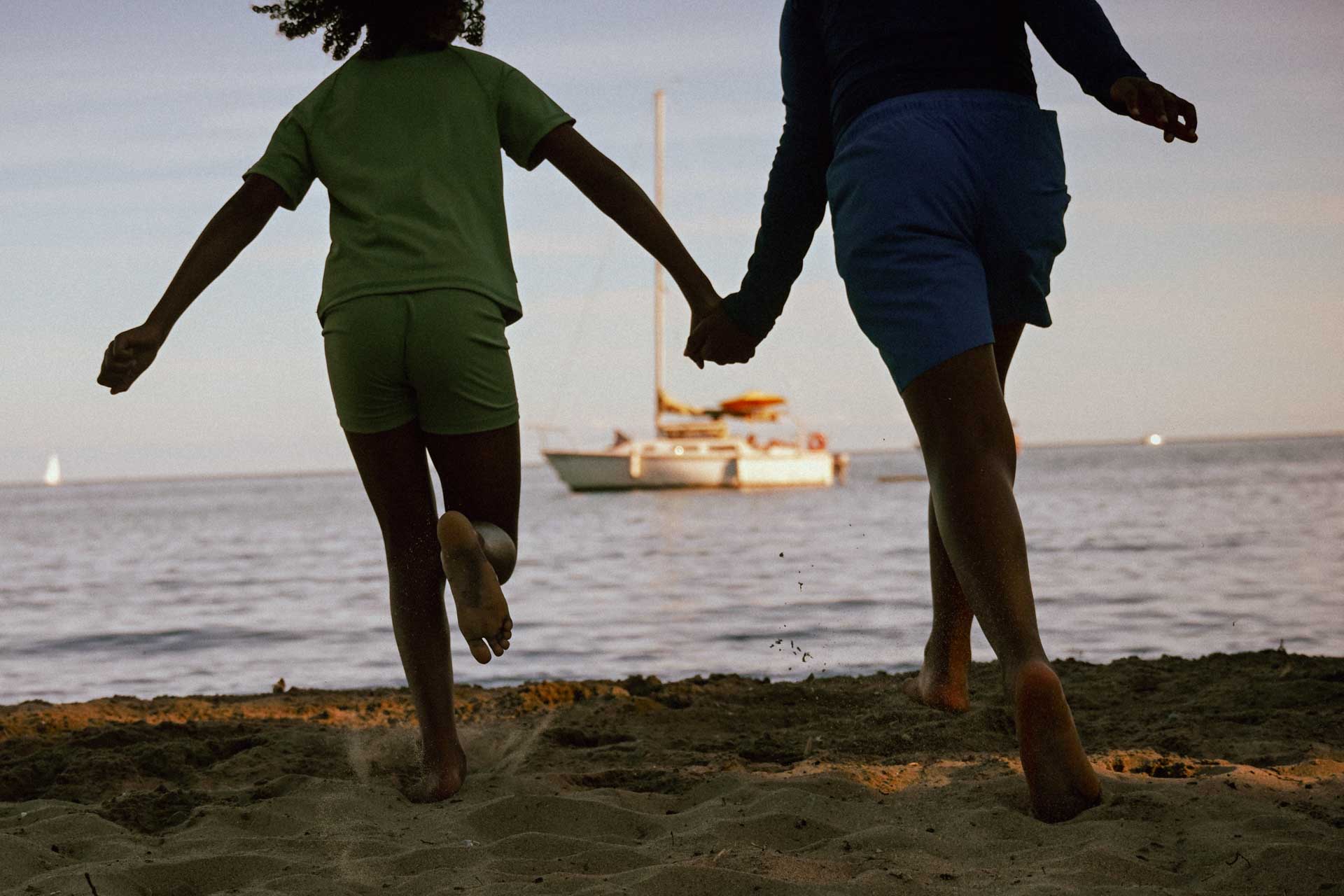 Two people holding hands run across a sandy beach towards the sea, with a boat offshore.