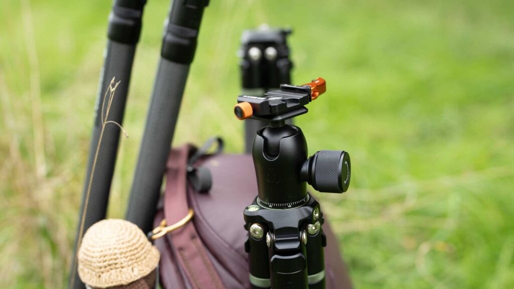 A ball head with an orange-accented quick-release clamp mounted on a tripod, with a backpack behind it.