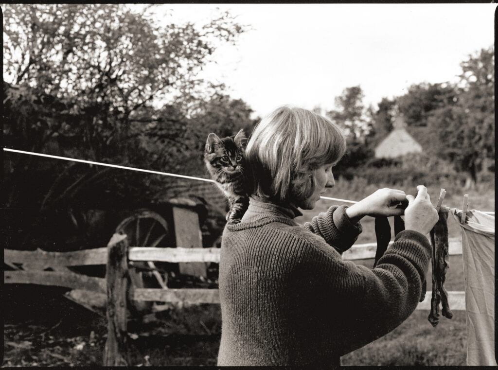 Sepia photograph of a woman pegging laundry onto a washing line outdoors, with a tabby kitten perched on her shoulder.