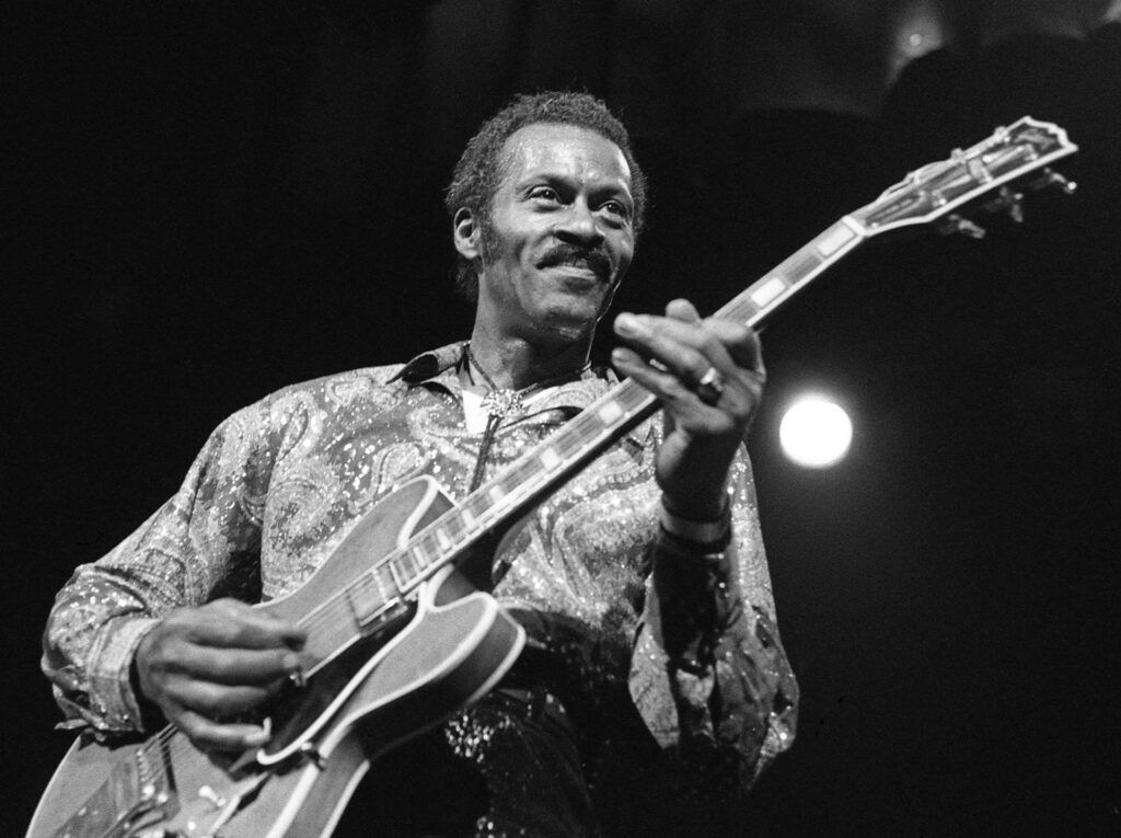 Black-and-white photograph of Chuck Berry smiling as he plays an electric guitar on stage, wearing a patterned shirt.