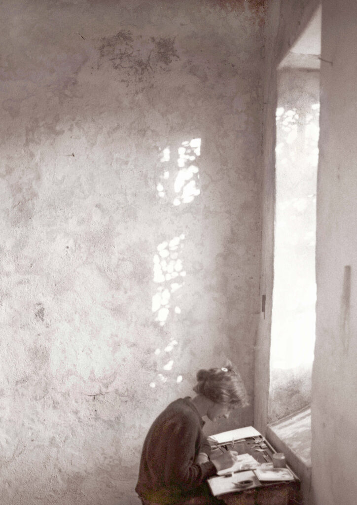 Sepia photograph of a woman writing at a small desk beside a tall window, with patches of sunlight falling across the textured wall.
