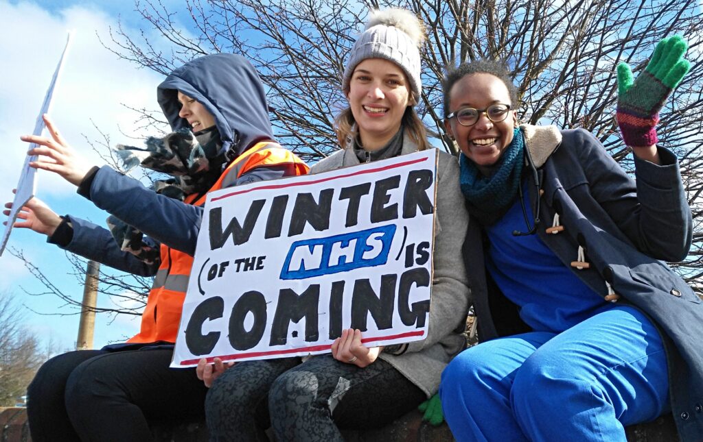 Three women outdoors at an NHS protest, one waving in blue scrubs and another holding a placard reading “Winter (of the NHS) is coming”.