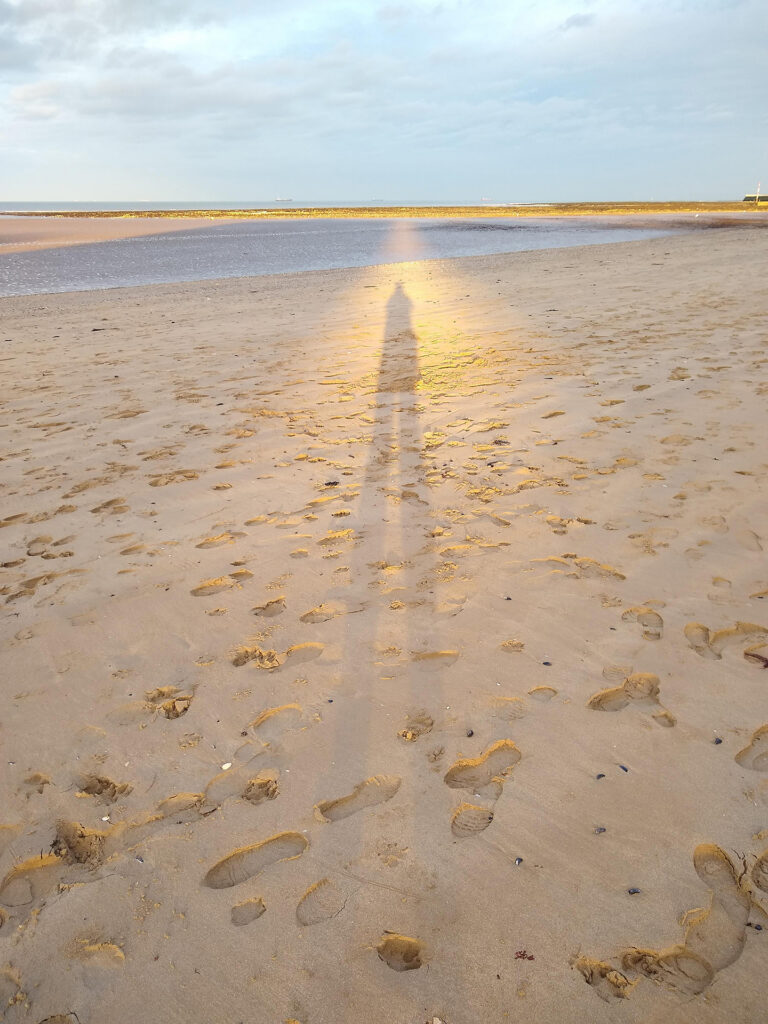 Sarah Wyld’s long shadow stretches across a sandy beach, surrounded by a golden glow, with footprints, a tidal pool and the sea beyond.